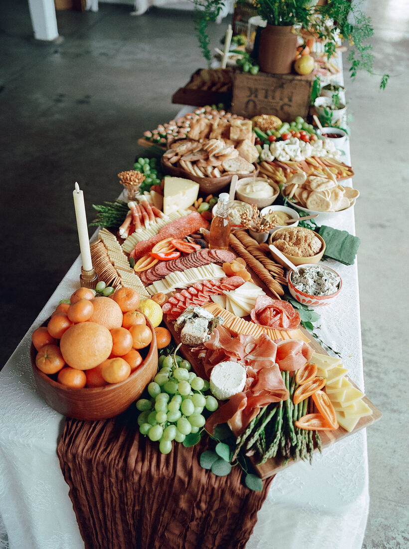 Long grazing table layered with cheeses, charcuterie, fruit, crudité and fresh flowers