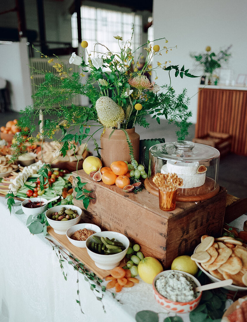 Grazing table styled with citrus, figs, olives, cheese under glass and wildflower arrangements