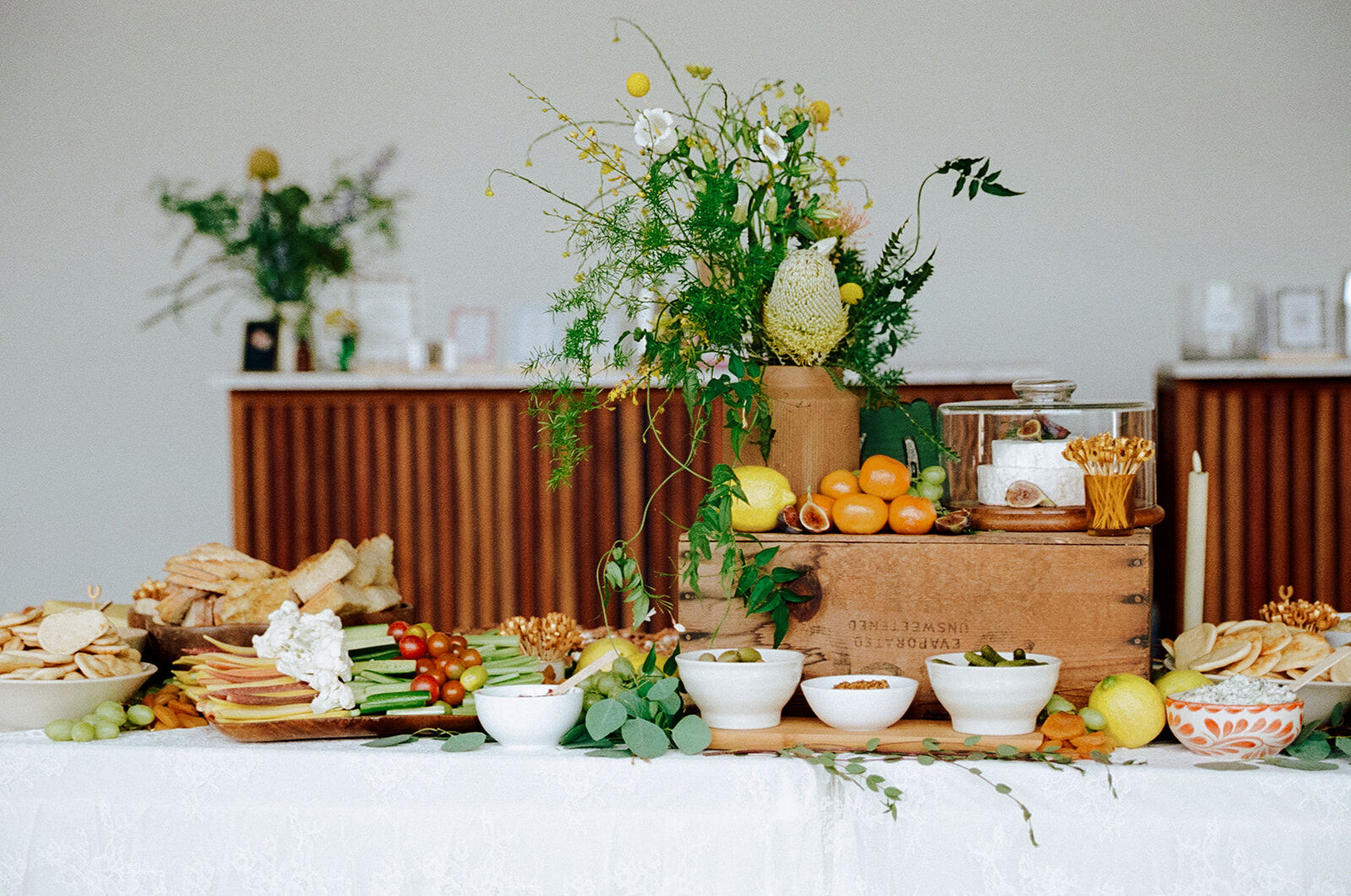 Chef-styled grazing table with wooden crates, wildflowers, cheese under a glass cloche and bowls of house accompaniments
