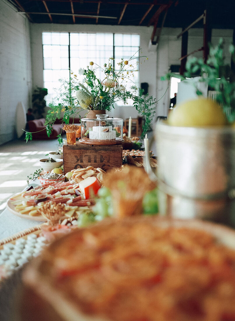 Grazing table with flowers and a cheese cloche in a sunlit warehouse venue